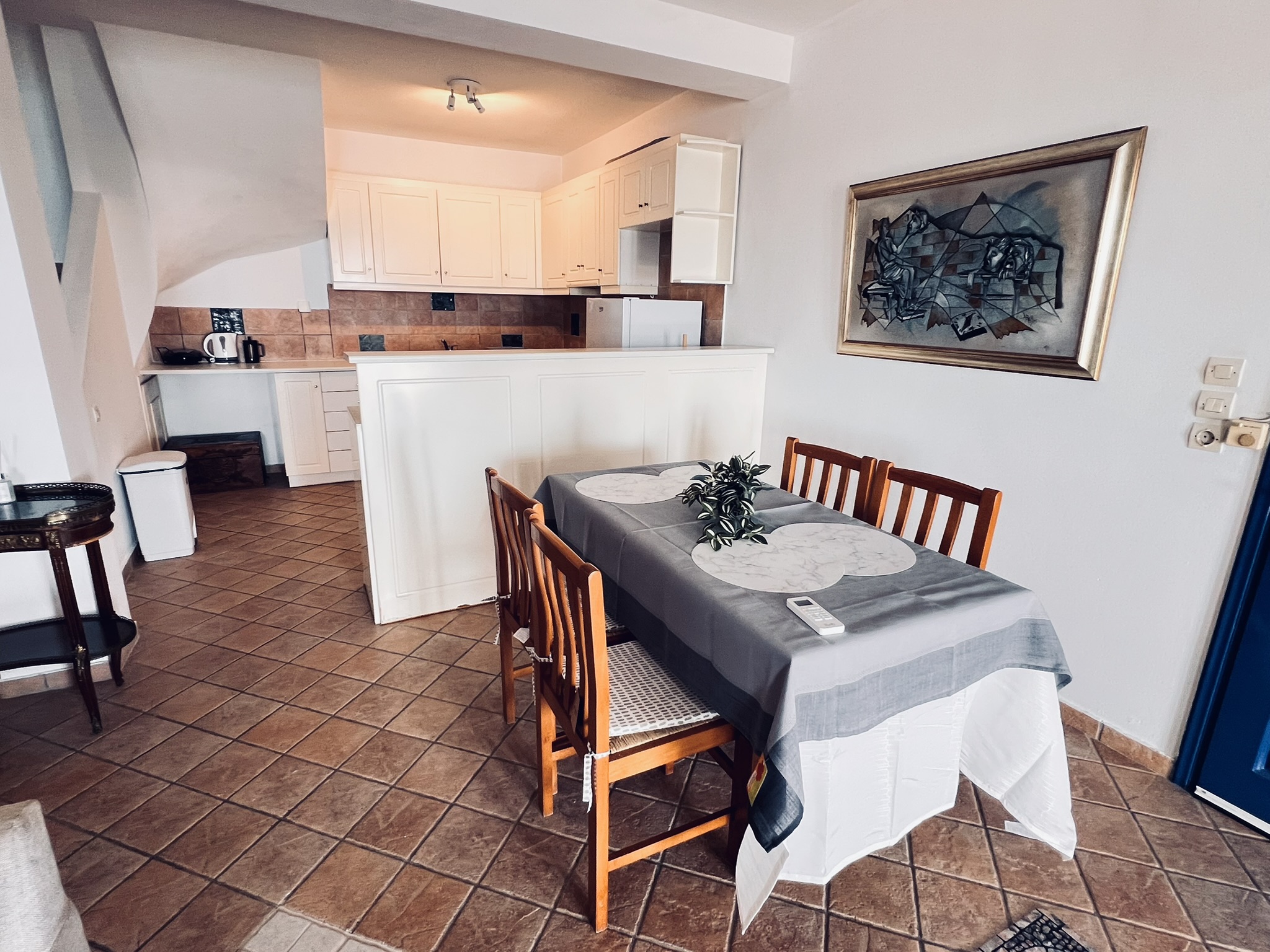 The dining table for four in front of the open kitchen — white cabinets, terracotta floor tiles, and framed painting above