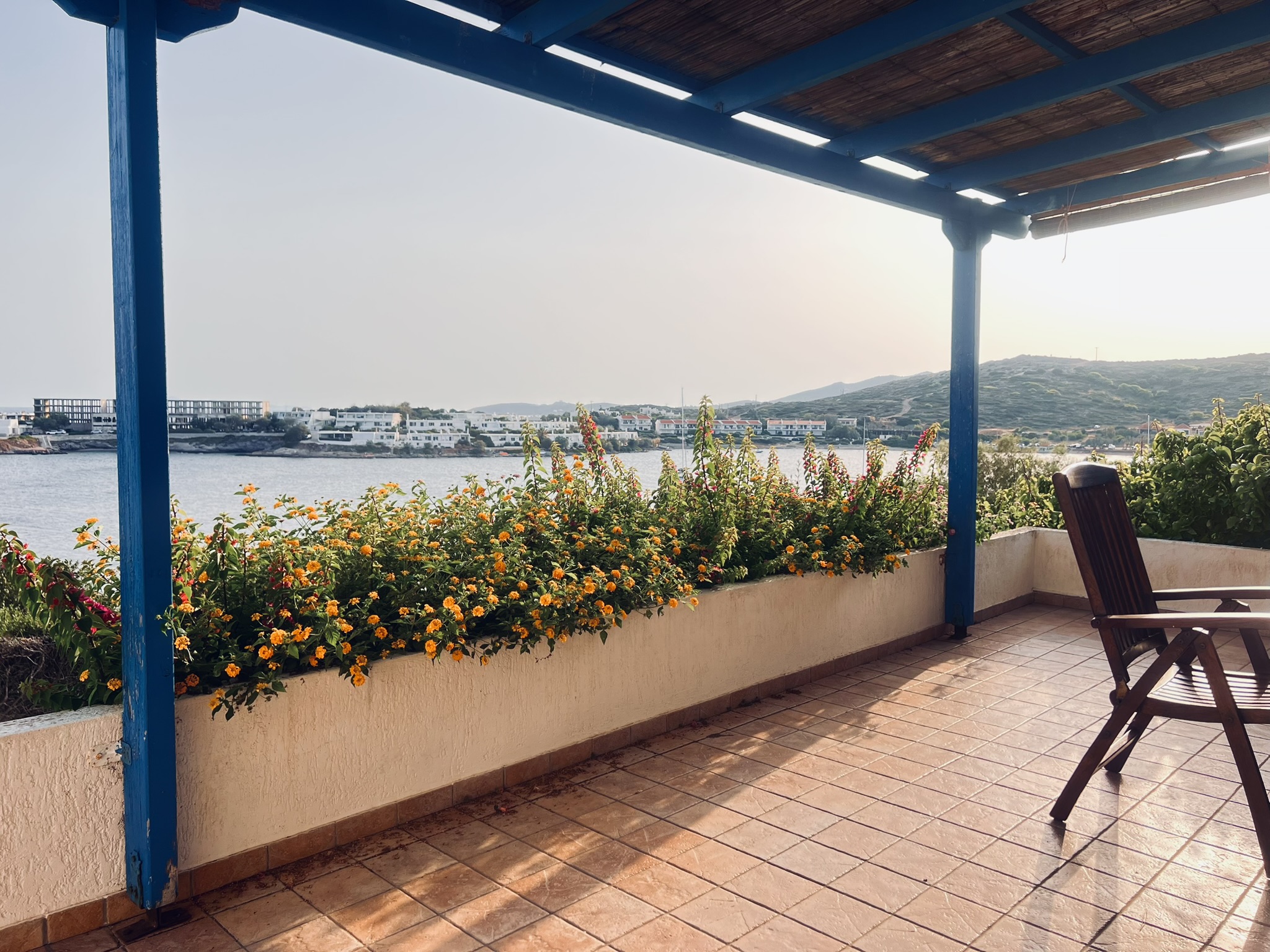 View from the terrace at Pounta Zeza — blue pergola, terracotta tiles, flowers, and the Aegean bay beyond